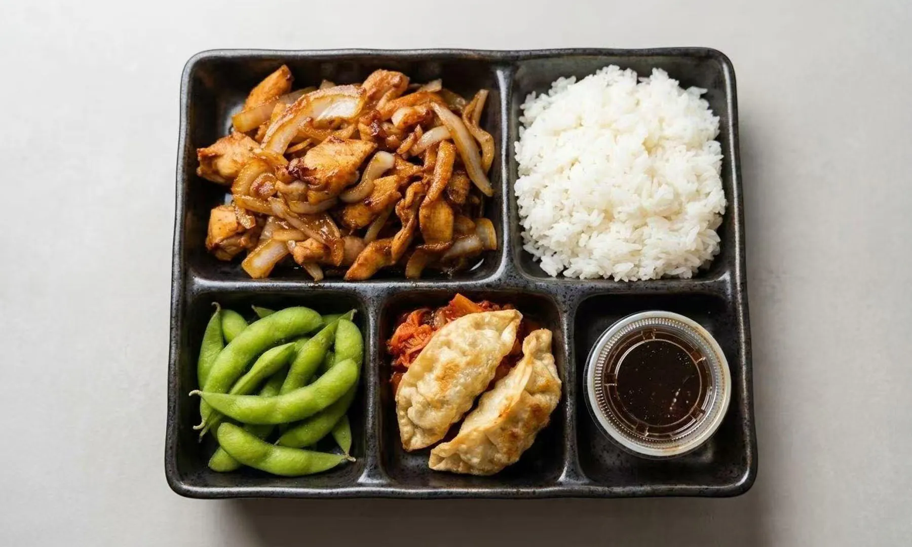 Bento box with teriyaki chicken, rice, edamame, gyoza, and kimchi