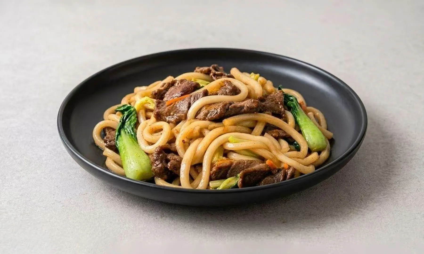 Stir-fried beef udon noodles with bok choy on a black plate